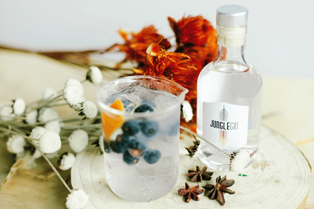 Garrafa de Jungle Gin ao lado de flores vermelhas e brancas e um copo de drink com frutas.
