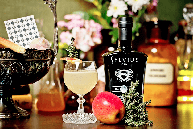 Garrafa do gin Sylvius ao lado de uma taça, uma maçã e uma cesta de metal. Ao fundo, várias garrafas e tubos de ensaio desfocadas.