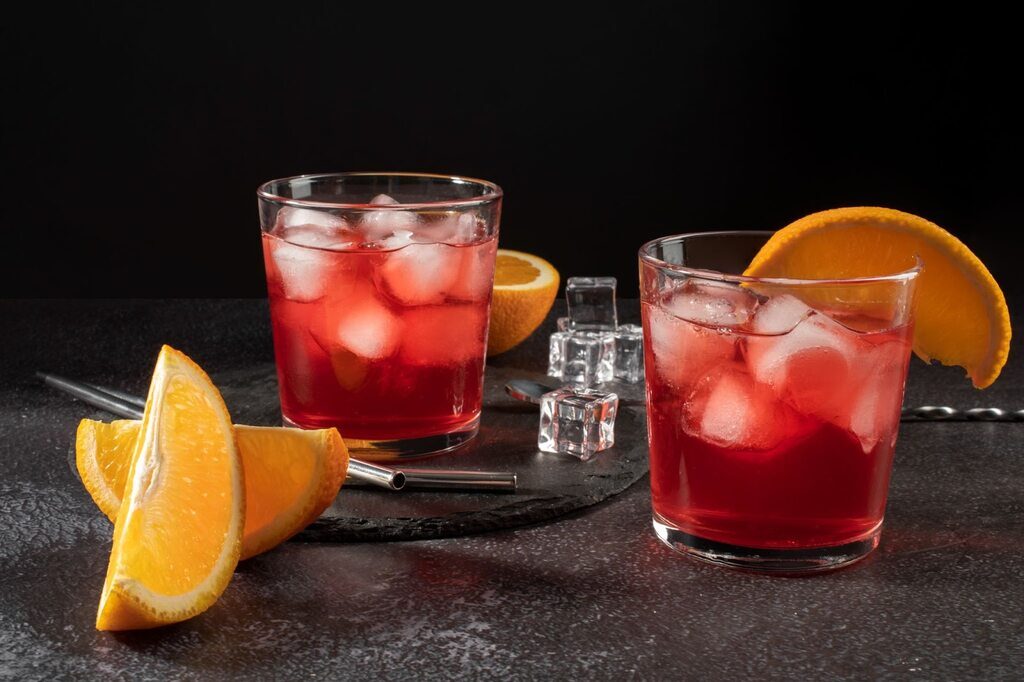 Dois copos servidos com o drink Negroni, um deles já está servido com uma rodela de laranja.