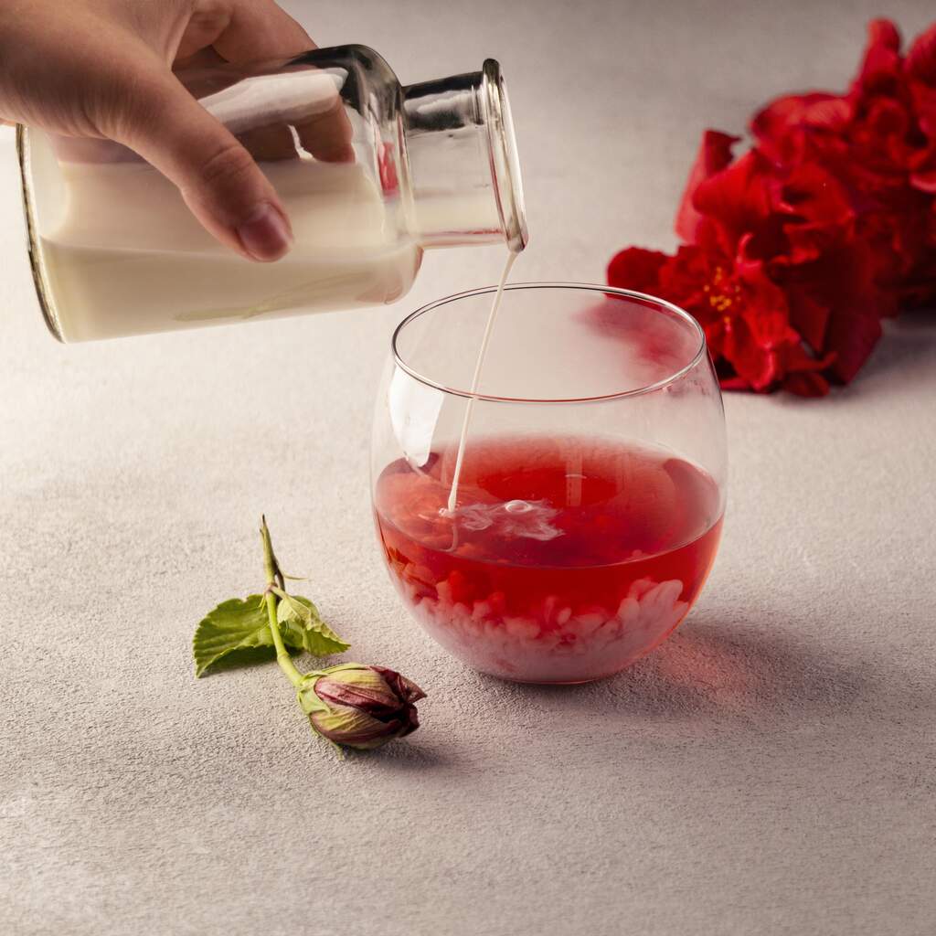 Copo de vidro servido com gin com Monin de morango e chá de hibisco, enquanto uma mão adiciona mais gin ao copo.