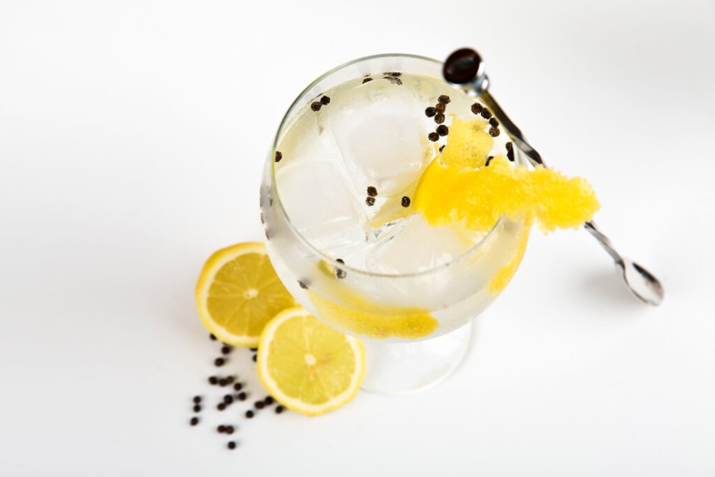 uma visão superior de uma taça  com drink de bebida destilada com pedaços de limão na mesa em que  mesma está com casca de limão na borda do copo e com uma colher escorada no mesmo e com cravos na bebida e na mesa