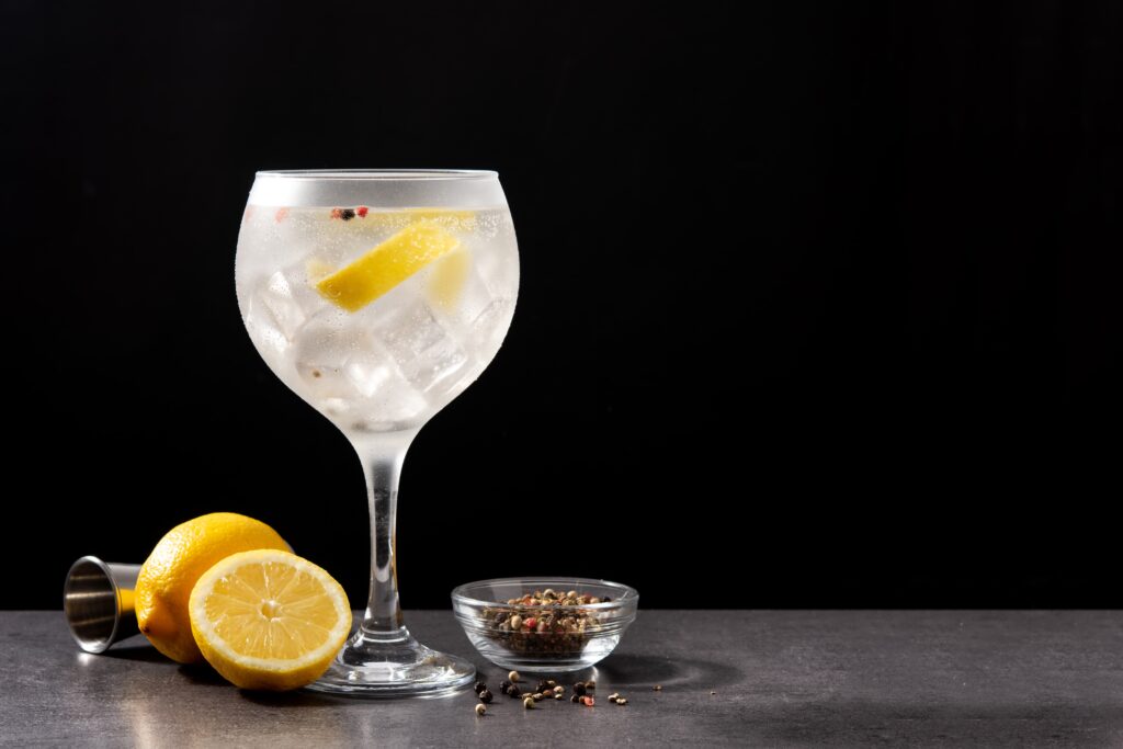 bebidas para eventos: Taça de gin com casca de limão dentro, com limões sobre uma mesa de mármore com cravos e sementes dentro de um pote de vidro