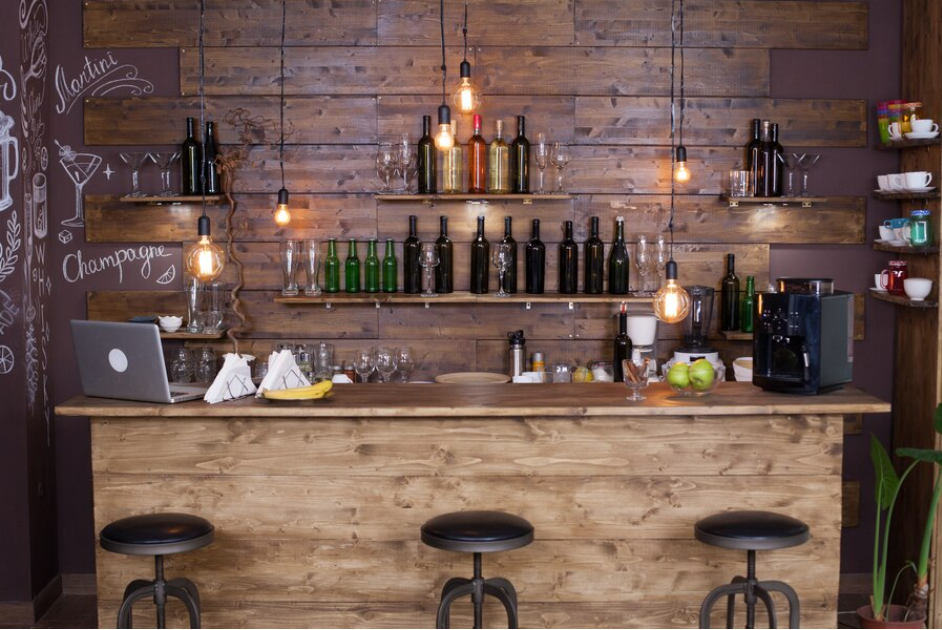 home bar: bar ornamentado em madeira, muitas garrafas de bebidas na prateleira de madeira, luzes vintage, na bancada do bar há frutas, um computador e guardanapos.