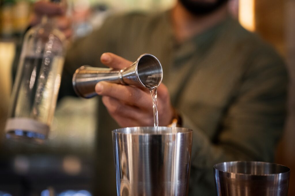 Montar um bar: Barman preparando um drink no mixer colocando uma dose de bebida alcoólica, ele está vestido com uma camisa cinza e com uma garrafa de bebida em sua mão direita
